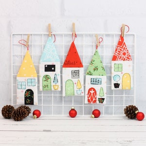 Puede incluir: Cinco adornos navideños de fieltro coloridos con forma de casas con diferentes diseños y detalles. Cada casa tiene un fondo blanco con un techo rojo, verde, azul o amarillo. Las casas están colgadas de una rejilla de alambre blanca.
