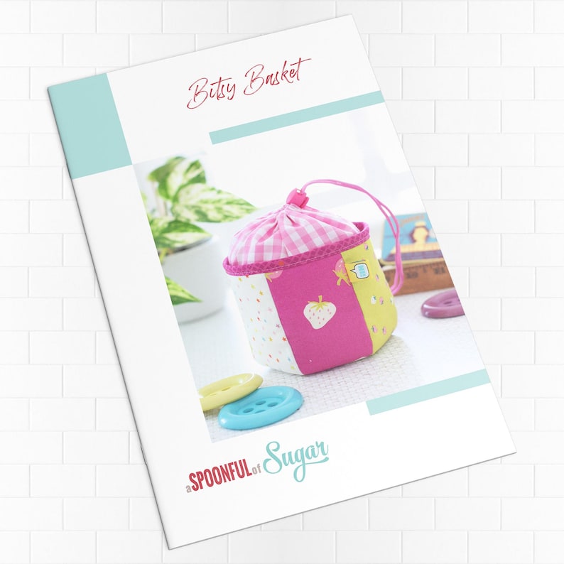Pode incluir: Um padr&atilde;o de costura intitulado "Bitsy Basket" com uma cesta colorida feita &agrave; m&atilde;o. A cesta apresenta pain&eacute;is de tecido rosa, amarelo e branco, um topo xadrez e uma aplica&ccedil;&atilde;o de morango. V&aacute;rios bot&otilde;es em tons de azul e amarelo est&atilde;o espalhados.