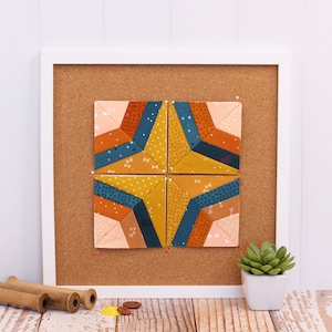 May include: A corkboard with a white frame, displaying a colorful patchwork quilt block with geometric shapes in shades of orange, yellow, blue, and pink. The quilt block is pinned to the corkboard with red push pins.