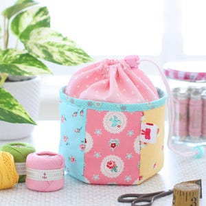 Pode incluir: Uma bolsa com cord&atilde;o de patchwork com uma parte superior rosa com bolinhas e um corpo com estampa floral em azul, rosa e amarelo. A bolsa est&aacute; ao lado de carret&eacute;is de linha, tesouras e uma fita m&eacute;trica. Uma pequena etiqueta com um la&ccedil;o vermelho est&aacute; anexada.