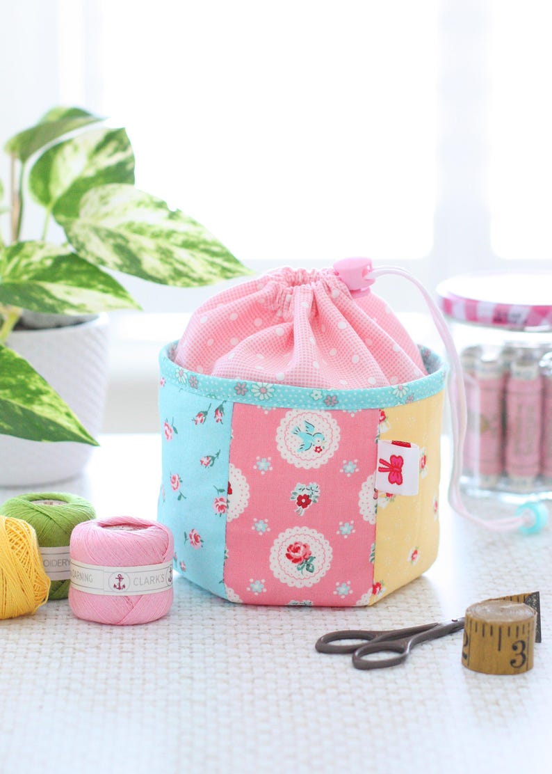 Pode incluir: Uma bolsa com cord&atilde;o de patchwork com uma parte superior rosa com bolinhas e um corpo com estampa floral em azul, rosa e amarelo. A bolsa est&aacute; ao lado de carret&eacute;is de linha, tesouras e uma fita m&eacute;trica. Uma pequena etiqueta com um la&ccedil;o vermelho est&aacute; anexada.
