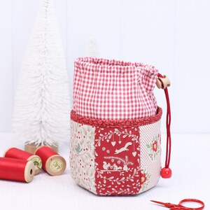 Pode incluir: Uma bolsa com cord&atilde;o de patchwork com uma parte superior xadrez vermelha e branca e uma base decorativa com padr&atilde;o vermelho e branco. A bolsa apresenta um cord&atilde;o vermelho com uma conta de madeira e uma conta vermelha. Tesouras vermelhas e carret&eacute;is de linha vermelha no fundo.