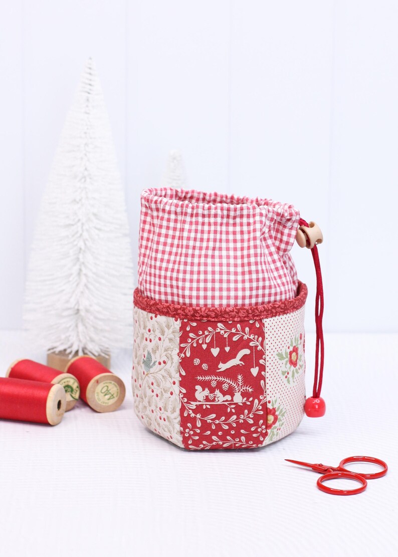 Pode incluir: Uma bolsa com cord&atilde;o de patchwork com uma parte superior xadrez vermelha e branca e uma base decorativa com padr&atilde;o vermelho e branco. A bolsa apresenta um cord&atilde;o vermelho com uma conta de madeira e uma conta vermelha. Tesouras vermelhas e carret&eacute;is de linha vermelha no fundo.