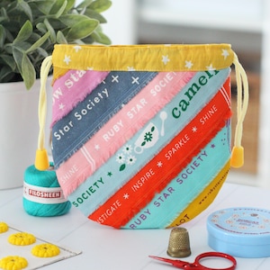 May include: A colorful drawstring bag with diagonal fabric strips, featuring text like "Star Society" and "Ruby Star Society." The bag has a yellow drawstring and trim, and is surrounded by sewing notions like thread, scissors, and a thimble.