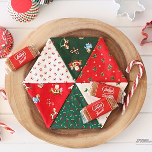 May include: A handmade Christmas-themed fabric coaster with a patchwork design. The coaster is surrounded by Lotus Biscoff cookies, candy canes, and festive cupcake liners. The coaster features red, green, and white fabrics with holiday patterns.