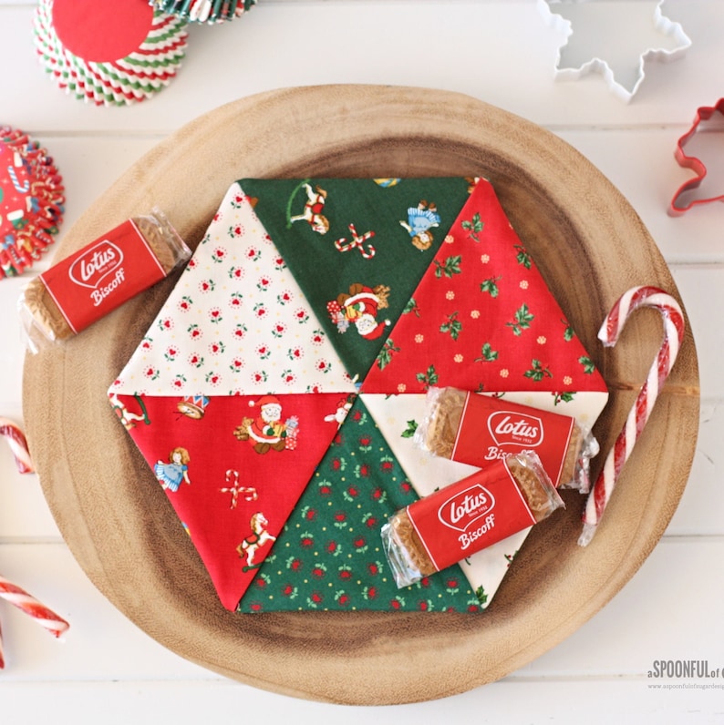 May include: A handmade Christmas-themed fabric coaster with a patchwork design. The coaster is surrounded by Lotus Biscoff cookies, candy canes, and festive cupcake liners. The coaster features red, green, and white fabrics with holiday patterns.