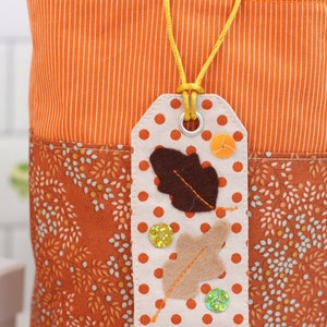 K&ouml;nnte beinhalten: Eine wei&szlig;e Stofftasche mit einem orange-wei&szlig; gestreiften Stoffoberteil und einem braun-wei&szlig; gemusterten Stoffunterteil. Ein wei&szlig;es Stoff-Etikett mit braunen und braunen Filzbl&auml;ttern, orangefarbenen und gr&uuml;nen Glitzerakzenten und wei&szlig;en Punkten ist mit einer gelben Schnur an der Tasche befestigt.