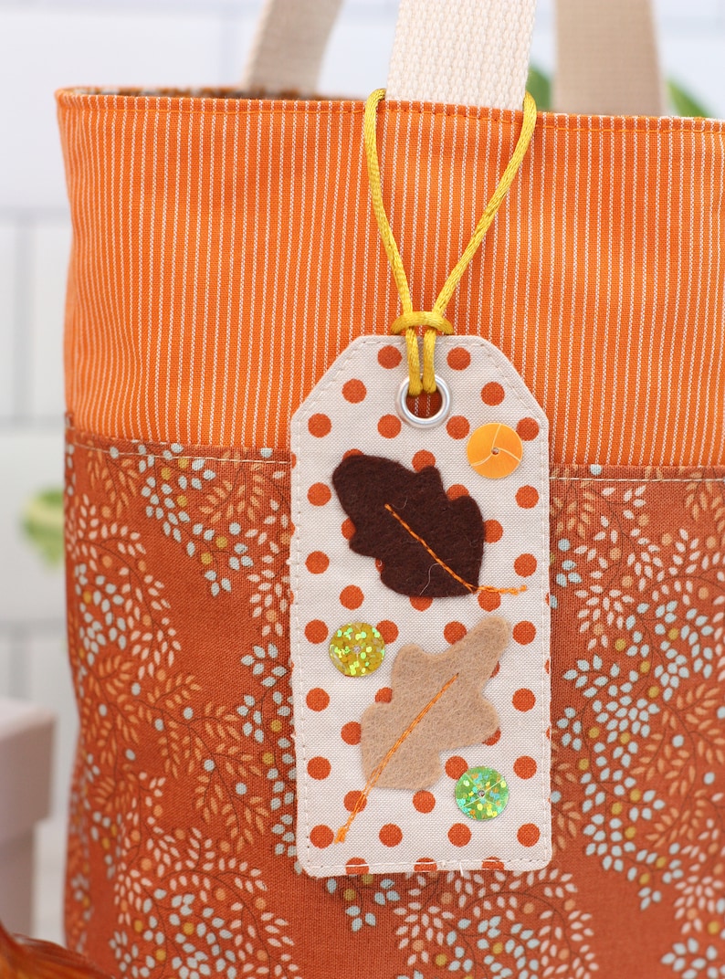 K&ouml;nnte beinhalten: Eine wei&szlig;e Stofftasche mit einem orange-wei&szlig; gestreiften Stoffoberteil und einem braun-wei&szlig; gemusterten Stoffunterteil. Ein wei&szlig;es Stoff-Etikett mit braunen und braunen Filzbl&auml;ttern, orangefarbenen und gr&uuml;nen Glitzerakzenten und wei&szlig;en Punkten ist mit einer gelben Schnur an der Tasche befestigt.
