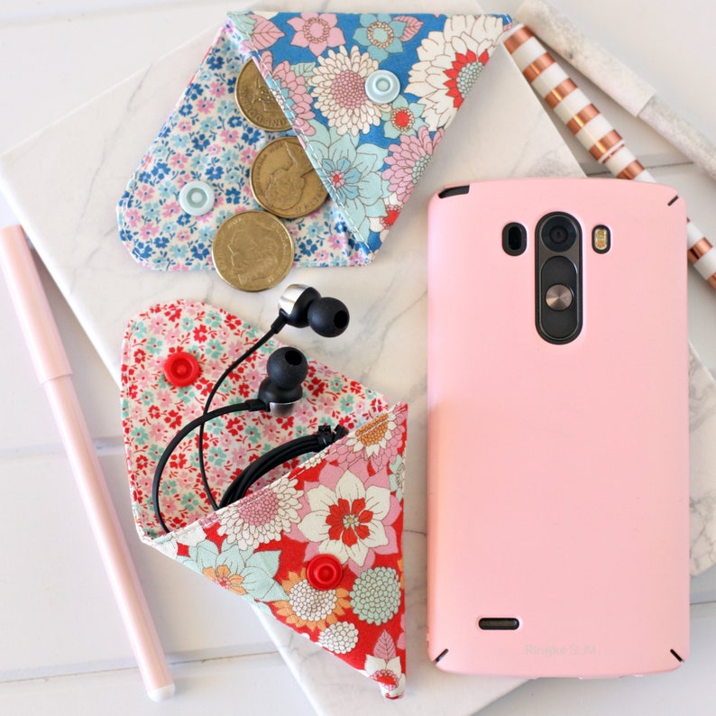 K&ouml;nnte beinhalten: Zwei kleine Stoffbeutel mit Blumenmustern. Ein Beutel enth&auml;lt M&uuml;nzen und der andere ein Paar schwarze Ohrh&ouml;rer. Ein rosa Smartphone ist ebenfalls auf dem Bild zu sehen.