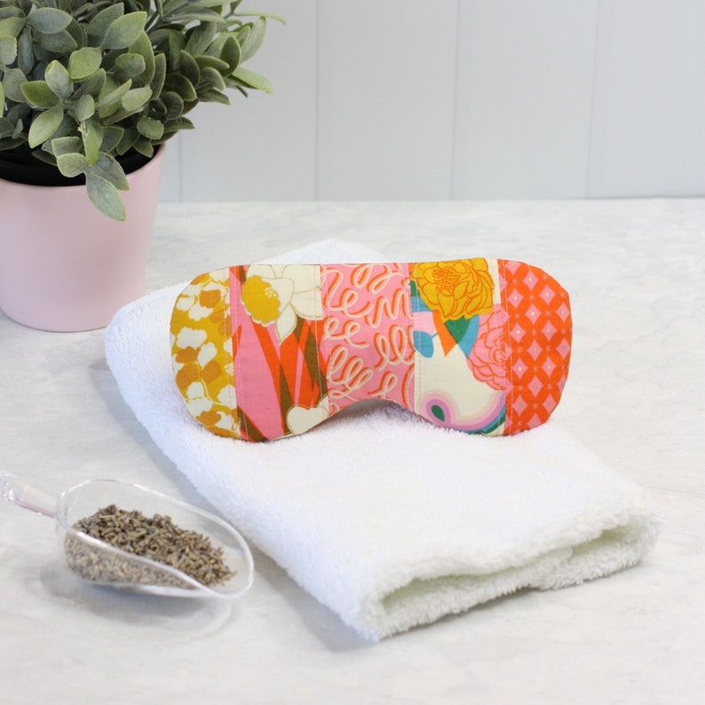May include: A colorful patchwork eye mask with pink, orange, yellow, and green floral patterns. The eye mask is lying on a white towel.