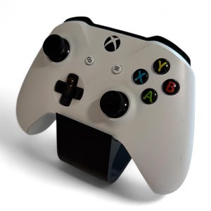 May include: A white Xbox controller with black joysticks, buttons, and a black support. The controller features the Xbox logo, a directional pad, and colored buttons (X, Y, A, B).