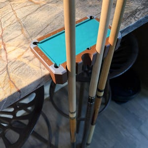 May include: A brown and gray plastic pool cue holder shaped like a miniature billiard table with a teal felt surface. The holder is designed to mount on the edge of a table and features three circular cutouts to securely hold three pool cues.