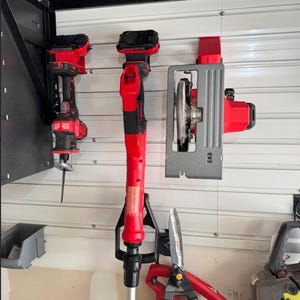 May include: A collection of red and black power tools, including a reciprocating saw, a string trimmer, and a circular saw, mounted on a white wall. Also visible are a pair of red Fiskars shears and a yellow and black chainsaw.