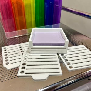 May include: A white, square desk organizer holding a stack of lavender sticky notes. Three white, slotted, rectangular dividers are arranged around the organizer. A clear plastic container filled with rainbow-colored file folders is in the background.