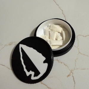 May include: Open black container with a white interior, filled with white pouches. The lid, placed to the side, features a white arrowhead design on a black background. The container sits on a marble-like surface.