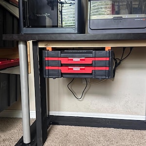 May include: A home workshop setup featuring a 3D printer and an additional printer. Below the desk are two black and red storage drawers. The setup is on a beige carpet.