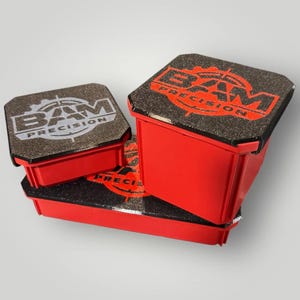May include: Three red plastic storage containers with black textured lids. Each lid features the text BAM PRECISION printed in a bold, stylized font with a target graphic. The containers are arranged in a stack, showing varying sizes and depths.