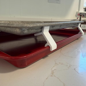 May include: A red baking sheet with a metal baking pan supported by white plastic brackets. The metal pan is a dull silver color. The brackets are designed to elevate the metal pan above the red baking sheet.