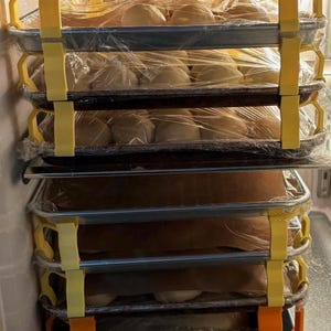 May include: A stack of baking trays filled with dough, wrapped in plastic, and stored in a refrigerator. The trays are held together by yellow and orange supports, creating a multi-tiered storage system for baked goods.