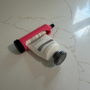 Puede incluir: Un exprimidor de tubos rosa con un mango negro está exprimiendo un tubo blanco de pasta de dientes. El tubo de pasta de dientes lleva la etiqueta "REFRESH MINT" y "Peso neto 141 g". El exprimidor está sobre una superficie blanca.