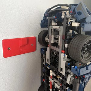 May include: A blue and black Lego Ford GT model car mounted on a white wall. The car is upside down, revealing its intricate internal structure and wheels. A red wall mount with the text "FORD GT" is attached to the wall.