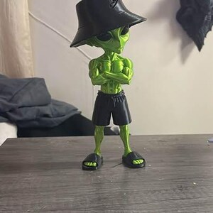 May include: A green alien figurine with a muscular build, wearing a black bucket hat, black shorts, and black slide sandals. The alien has its arms crossed over its chest and is standing in an upright position with a smooth, glossy finish.