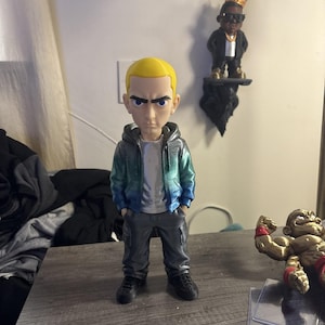 Puede incluir: Una figura estilizada de una persona con cabello corto amarillo y ojos azules. El personaje viste una sudadera con capucha plateada con un degradado azul y verde, una camiseta plateada, pantalones cargo gris oscuro y zapatos negros de pie.