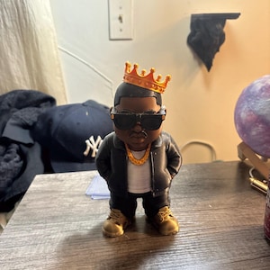 May include: A stylised figurine of a person wearing a black jacket, white shirt, and black trousers. The figure has dark skin, wears black sunglasses, a gold-coloured chain necklace, and a gold crown on their head. They are wearing tan-coloured boots.