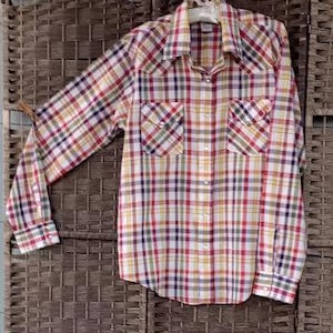 Puede incluir: Una camisa a cuadros de manga larga en una mezcla de colores que incluye rojo, amarillo, verde y azul. La camisa tiene dos bolsillos en el pecho y una botonadura. Está colgada de una percha blanca.