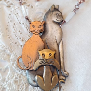 Puede incluir: Un collar de cobre, plata y oro con tres dijes de gato. Los gatos están apilados uno encima del otro, con el gato más grande en la parte inferior y el gato más pequeño en la parte superior. El collar está hecho de alambre de cobre y tiene cuentas blancas.