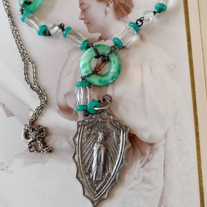 Könnte beinhalten: Eine antike grüne und silberne Halskette mit einem religiösen Anhänger, der eine Frau in einem langen Gewand zeigt. Der Anhänger trägt den Text "Ave Maria" und "Bitte für uns".