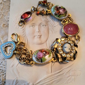 May include: A gold-toned bracelet with various charms and decorative elements. The bracelet features a heart-shaped charm with rhinestones, oval-shaped charms with painted floral designs, and a cameo pendant. The bracelet is displayed on a textured, off-white surface.