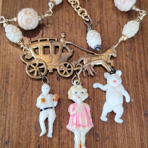 Puede incluir: Un collar de metal dorado con un dije de diligencia tirada por un caballo. Tres pequeños dijes cuelgan de la diligencia: una figura blanca, una niña con vestido rosa y un oso blanco. La cadena está decorada con cuentas talladas en crema y rosa.