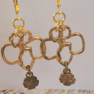 Può includere: Orecchini color oro con un design a forma di trifoglio, ognuno con una figura stilizzata e un piccolo ciondolo a forma di fiore. Gli orecchini hanno una chiusura a leva. Il design è intricato e dettagliato, con un'estetica vintage.