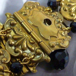 Könnte beinhalten: Eine goldfarbene Gürtelschnalle aus Metall mit aufwendigen floralen Designs und einem Scharnierverschluss. Die Schnalle ist mit schwarzen Glasperlen verziert.