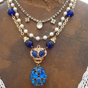 May include: A layered necklace with a blue and gold crown pendant and a blue crystal ball charm. The necklace features a pearl chain, a gold chain, and a rhinestone chain.