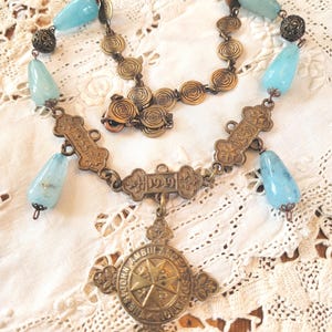 May include: Vintage necklace featuring a pendant with the text "ST JOHN AMBULANCE" and the year "1949". The necklace has light blue teardrop-shaped beads and ornate bronze-colored metalwork. The necklace is displayed on a white lace surface.