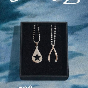Peut inclure: Deux colliers argentés dans un coffret noir. L'un a un pendentif en forme de goutte d'eau avec une étoile découpée, l'autre un porte-bonheur. L'image comprend le texte "Conan Gray" et "Wishbone" en écriture blanche sur fond bleu.