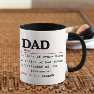 May include: White ceramic mug with a black handle and interior. The mug features the word "DAD" in large black letters, along with a definition that includes "fixer of everything" and "teller of bad jokes."