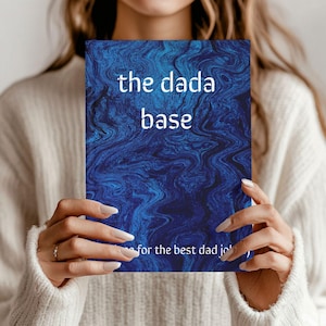 May include: A blue notebook with the text "the dada base" in white, held by a person. The notebook has a marbled design and the text "for the best dad jo" at the bottom. The person is wearing a cream-colored sweater.