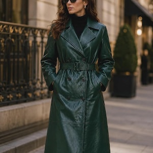 May include: A dark green, double-breasted trench coat featuring a wide collar, long sleeves, and a matching belt with a buckle at the waist. The coat has a smooth, polished finish and is worn over a black turtleneck sweater and dark sunglasses.