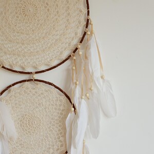 May include: Cream-colored crochet dream catcher with two circular frames. The frames are wrapped in brown material and adorned with white feathers, wooden beads, and cream-colored lace. A decorative wall hanging.