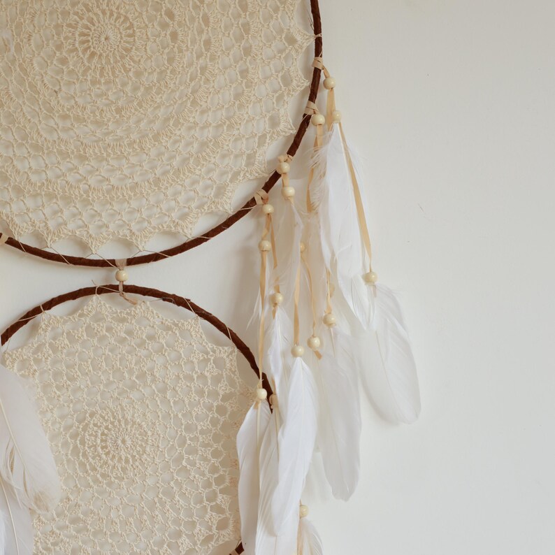 May include: Cream-colored crochet dream catcher with two circular frames. The frames are wrapped in brown material and adorned with white feathers, wooden beads, and cream-colored lace. A decorative wall hanging.