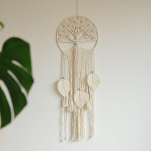 Op de afbeelding: Crèmekleurige macramé wandhanger met een levensboomontwerp in een cirkelvormig frame. Het stuk bevat hangende kwasten en drie bladvormige accenten, wat een bohemien touch toevoegt aan de interieurdecoratie.