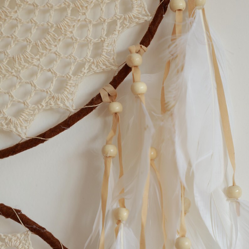 May include: Cream-colored dreamcatcher with a crocheted web design, brown branch accents, and dangling white feathers. Wooden beads are strung along the ribbons and branch. A decorative piece for a bedroom or living space.