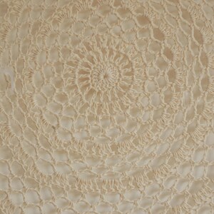 May include: Close-up of a cream-colored, handmade crochet doily. The circular design features concentric patterns of openwork and solid stitches, creating a delicate, textured surface. The central area has a tight spiral, radiating outwards.