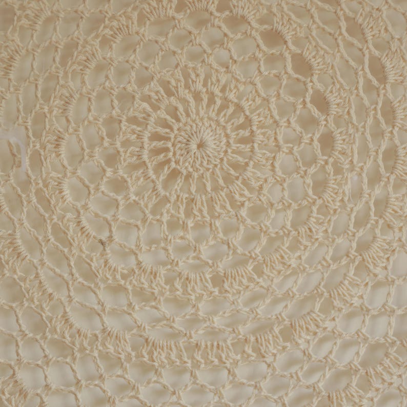 May include: Close-up of a cream-colored, handmade crochet doily. The circular design features concentric patterns of openwork and solid stitches, creating a delicate, textured surface. The central area has a tight spiral, radiating outwards.