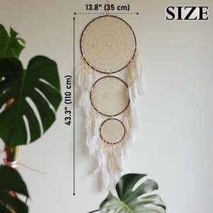 May include: A cream-colored dreamcatcher with three circular woven sections, adorned with white feathers and beads. The dreamcatcher is 43.3 inches (110 cm) long and 13.8 inches (35 cm) wide, designed for wall decor.
