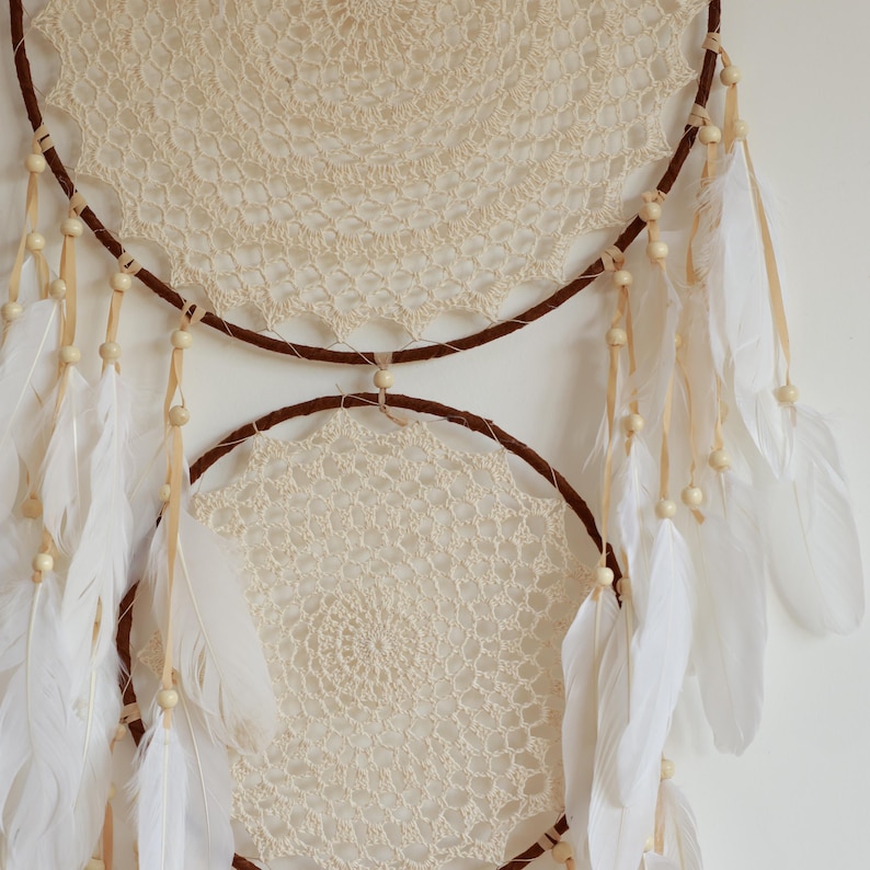 May include: Cream-colored dreamcatcher with two circular frames, adorned with a crocheted web design. Brown accents and white feathers hang from the bottom, with small wooden beads adding detail. A decorative piece.