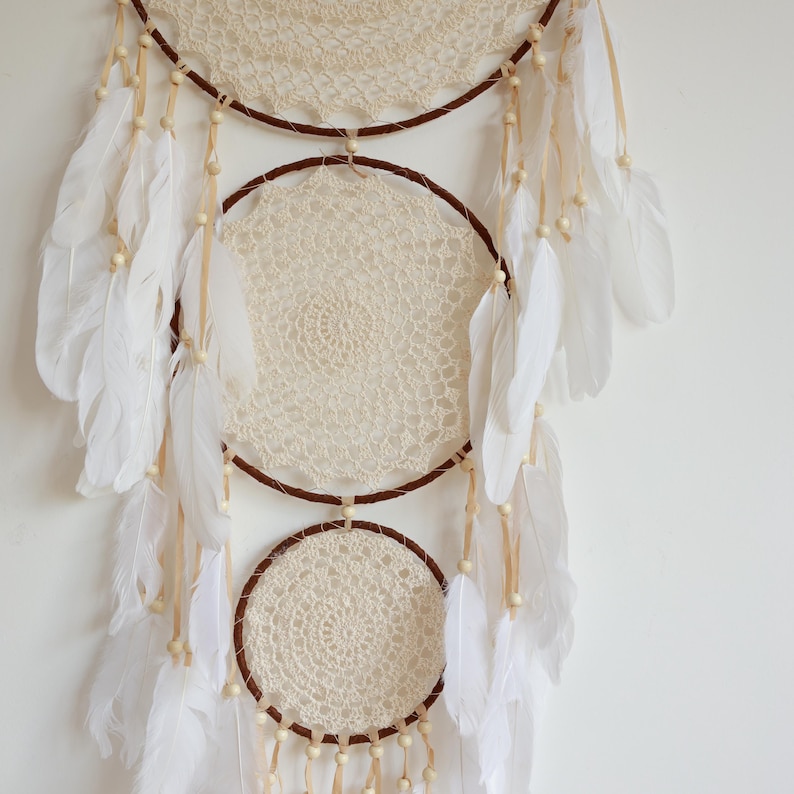May include: A handcrafted dreamcatcher with three circular frames. The frames are wrapped in brown material and adorned with cream-colored crocheted centers. White feathers and wooden beads hang from the frames, creating a bohemian aesthetic.